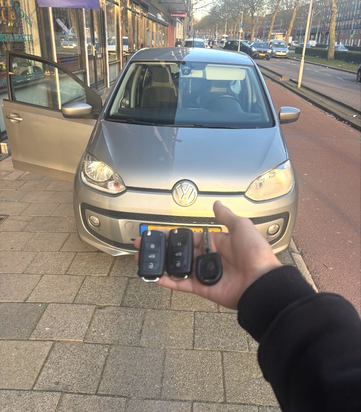 Drie autosleutels in een hand bij een zilverkleurige auto met open portier op straat